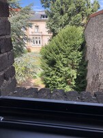 Vista a través de una ventana que muestra un edificio con un muro de piedra, un sendero y un árbol grande.
