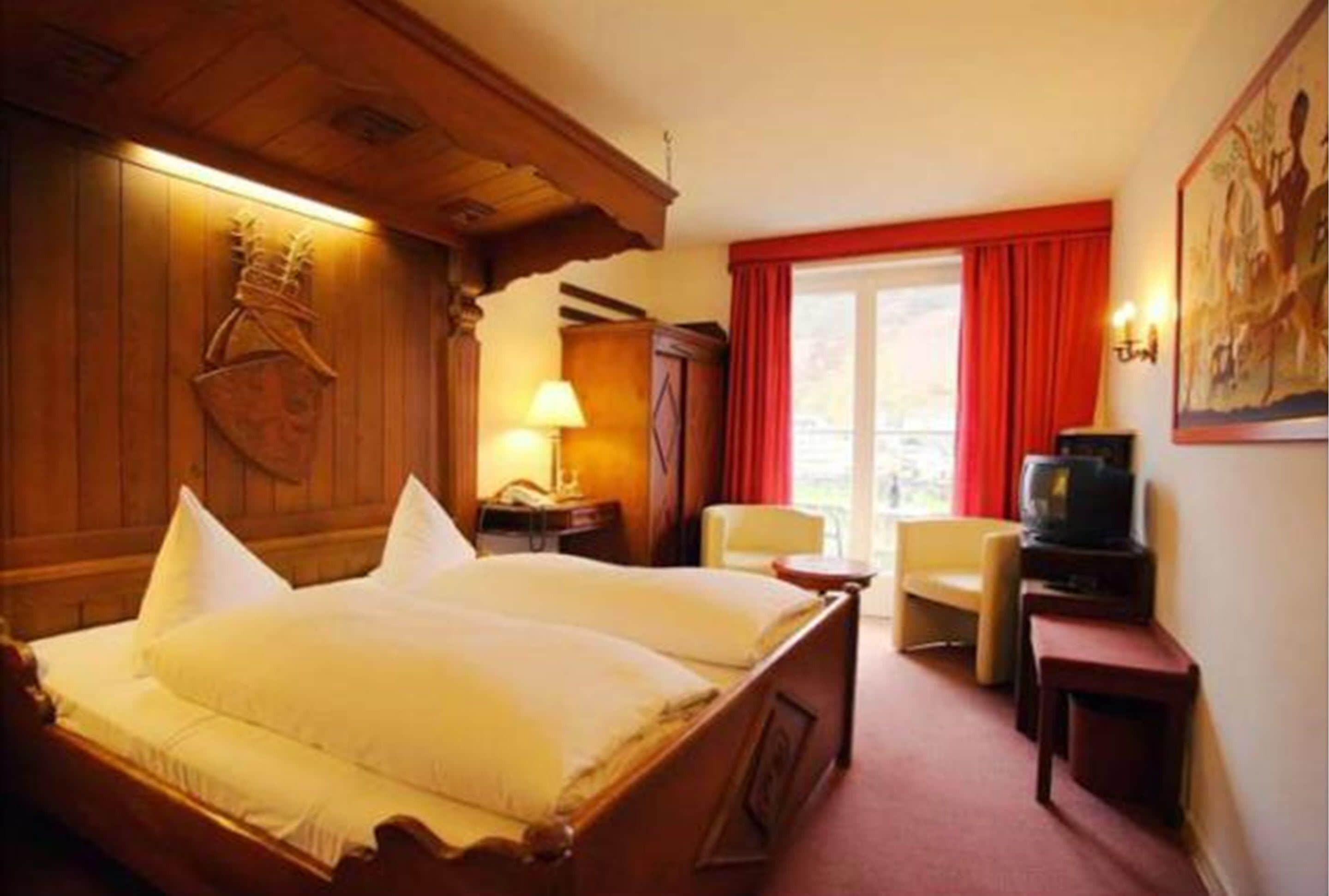 Una camera d'albergo con un letto di legno, una televisione, una sedia e una finestra con tende rosse.