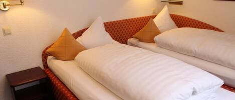 A hotel room with two beds, each with white linens and multiple pillows. There are wall-mounted lamps on either side of the beds.