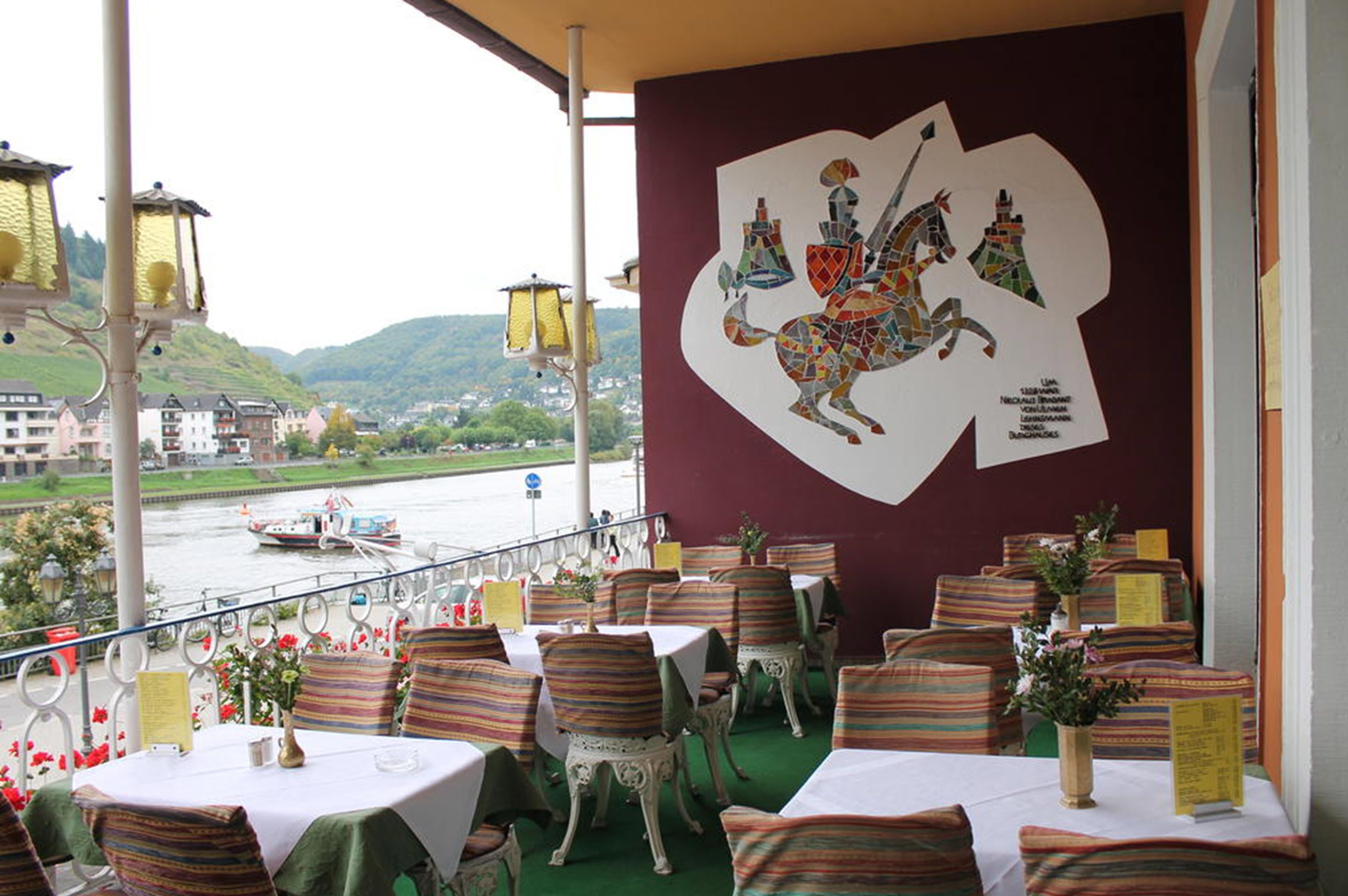 An outdoor dining area with tables and chairs, a view of a river, and a mural on the wall.