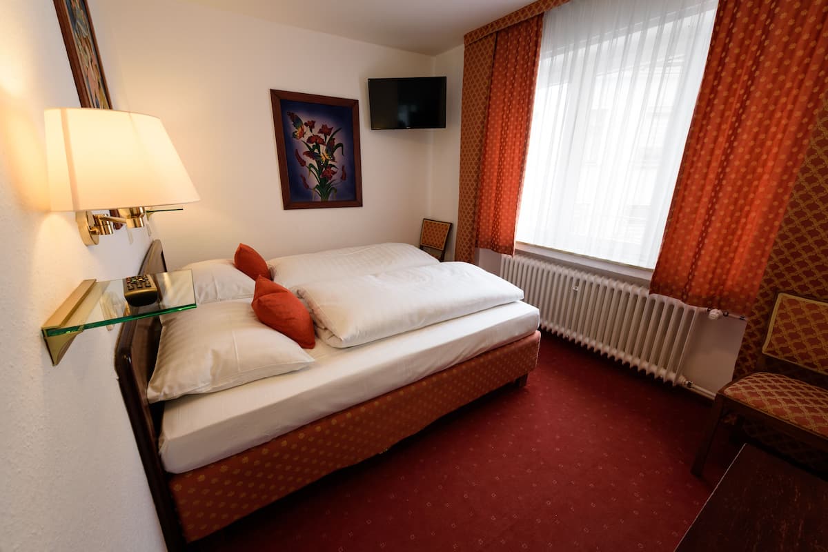 Habitación de hotel con cama, televisión, un cuadro, una silla y ventana con cortinas.