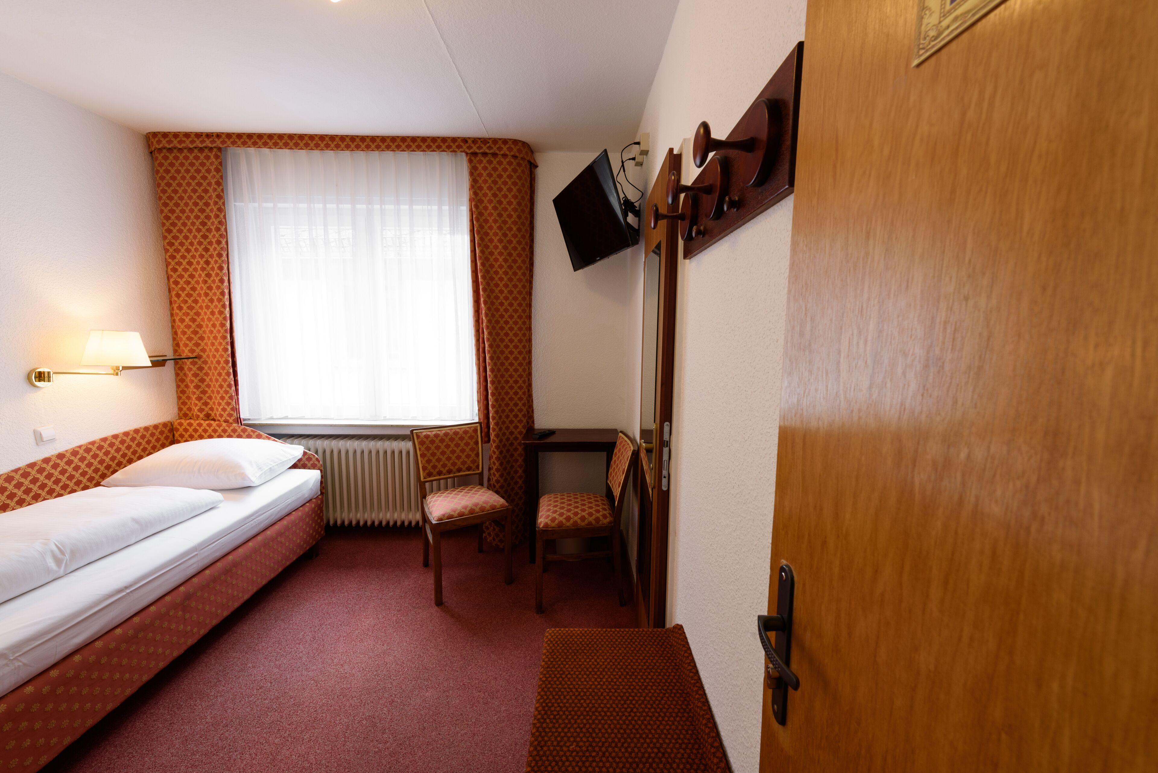 A small hotel room with a single bed, a desk with a chair, a TV mounted on the wall, and a radiator.