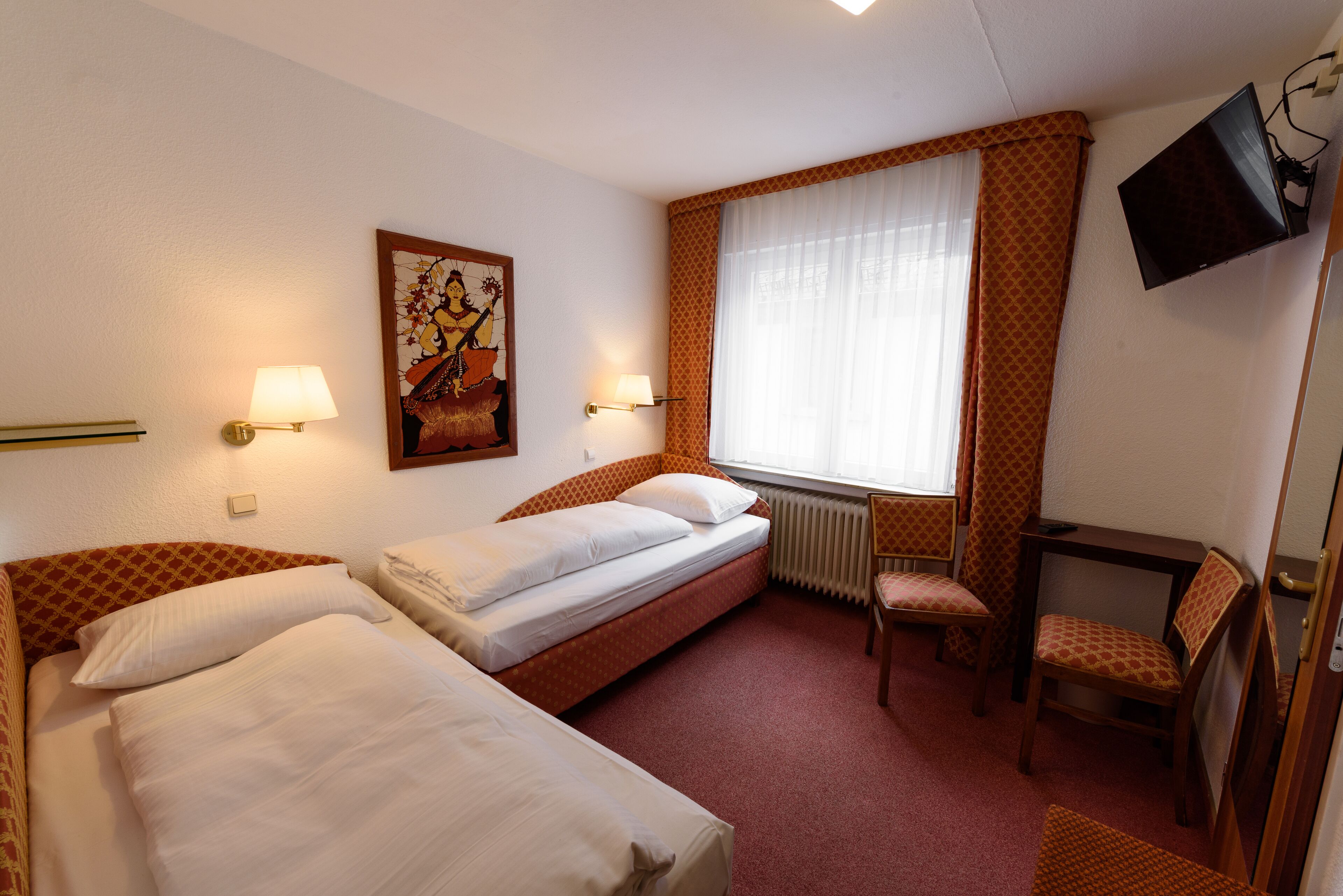 A hotel room with two beds, a television, a window with curtains, a radiator, and a chair.