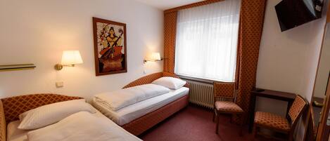 A hotel room with two beds, a television, a window with curtains, a radiator, and a chair.