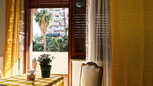 A room with a checkered table, a chair, a window with wooden shutters, and a view of buildings and trees outside. - A Casa di Frà B&B (Palermo)