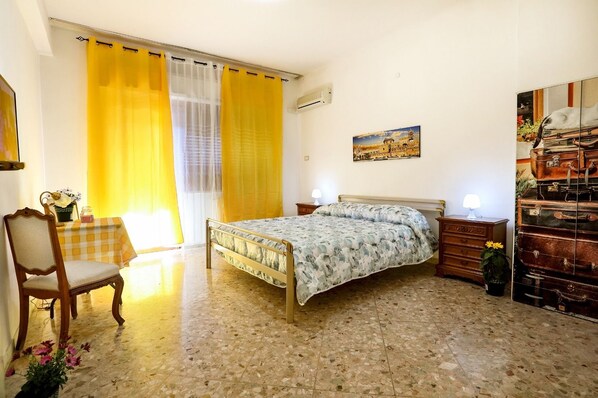 A bedroom with a bed, a chair, a table with a lamp, a painting, and a wall-mounted air conditioner.