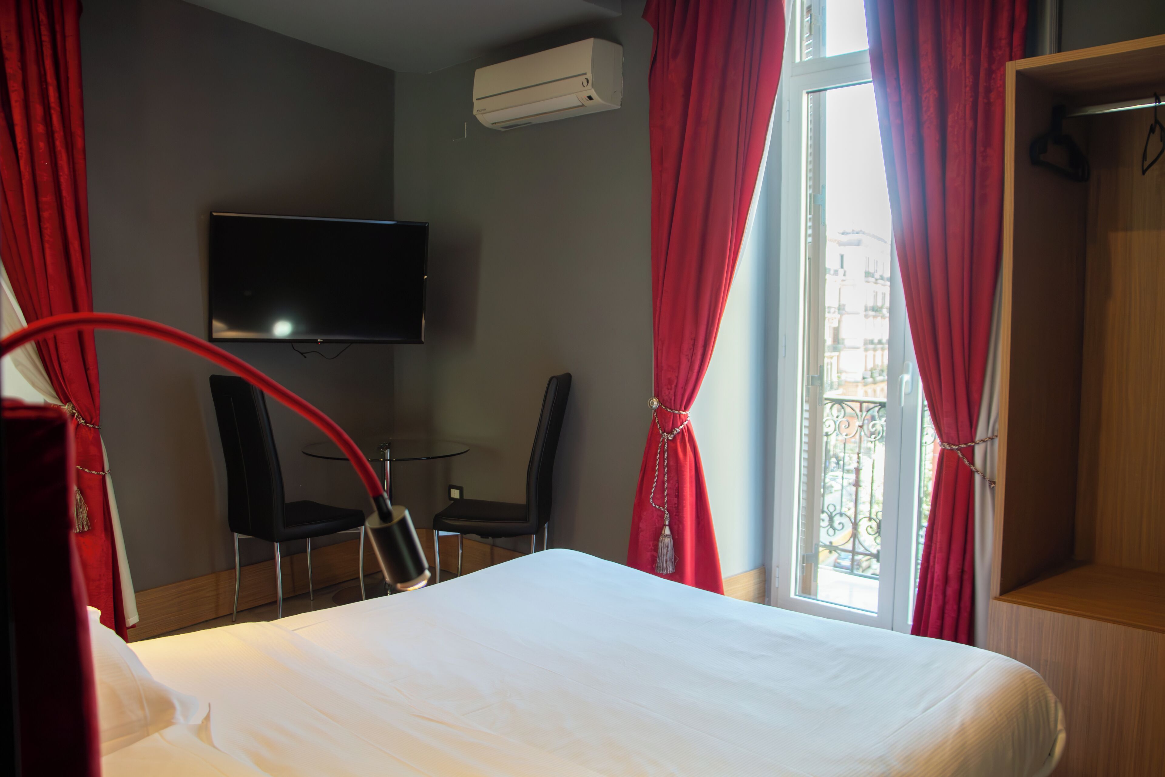 A hotel room with a bed, a TV mounted on the wall, a desk with a chair, and a balcony with red curtains.