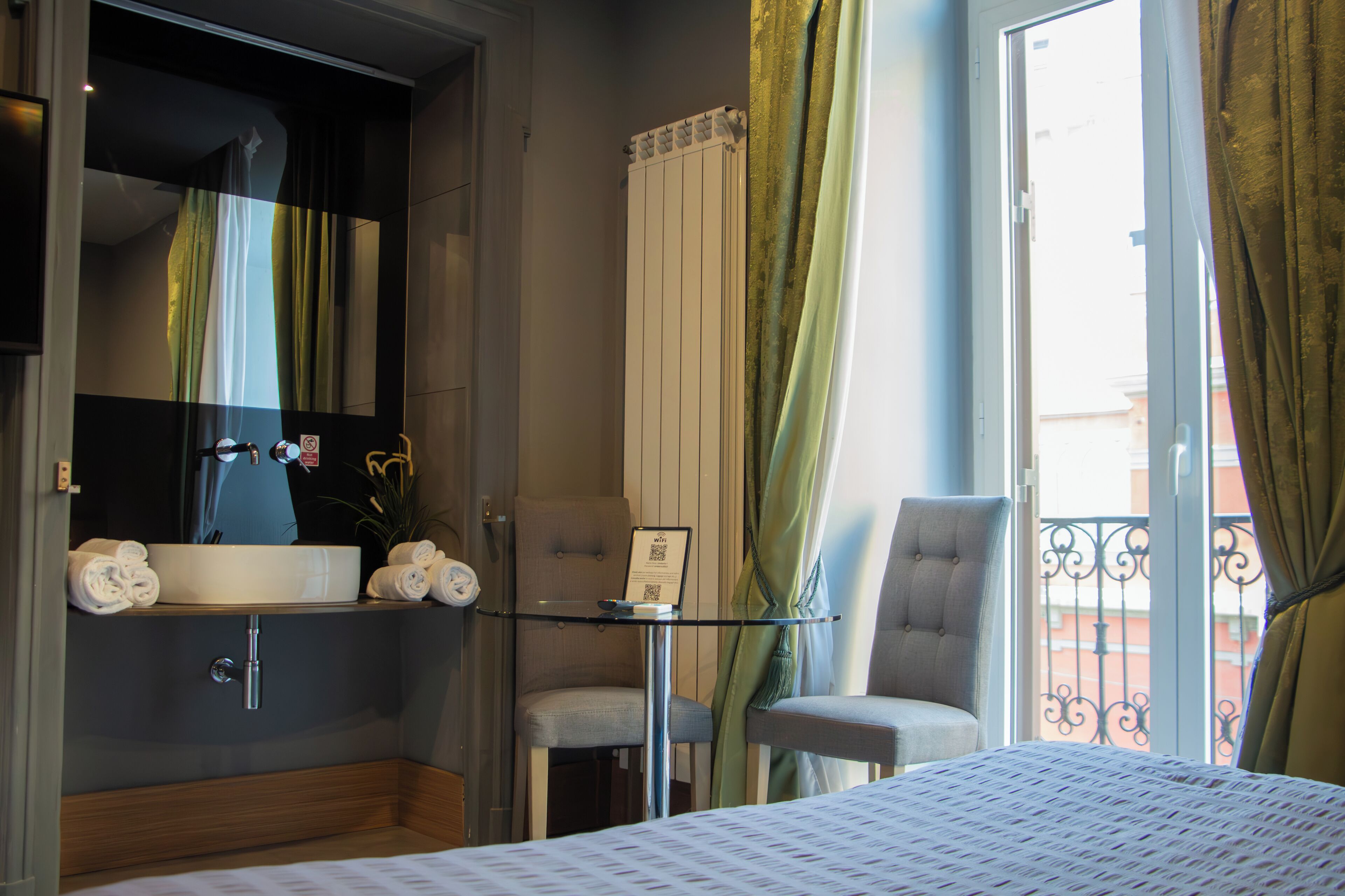 A hotel room with a large bed, a bedside table, a mirror, a sink, a chair, and a view of the outside.
