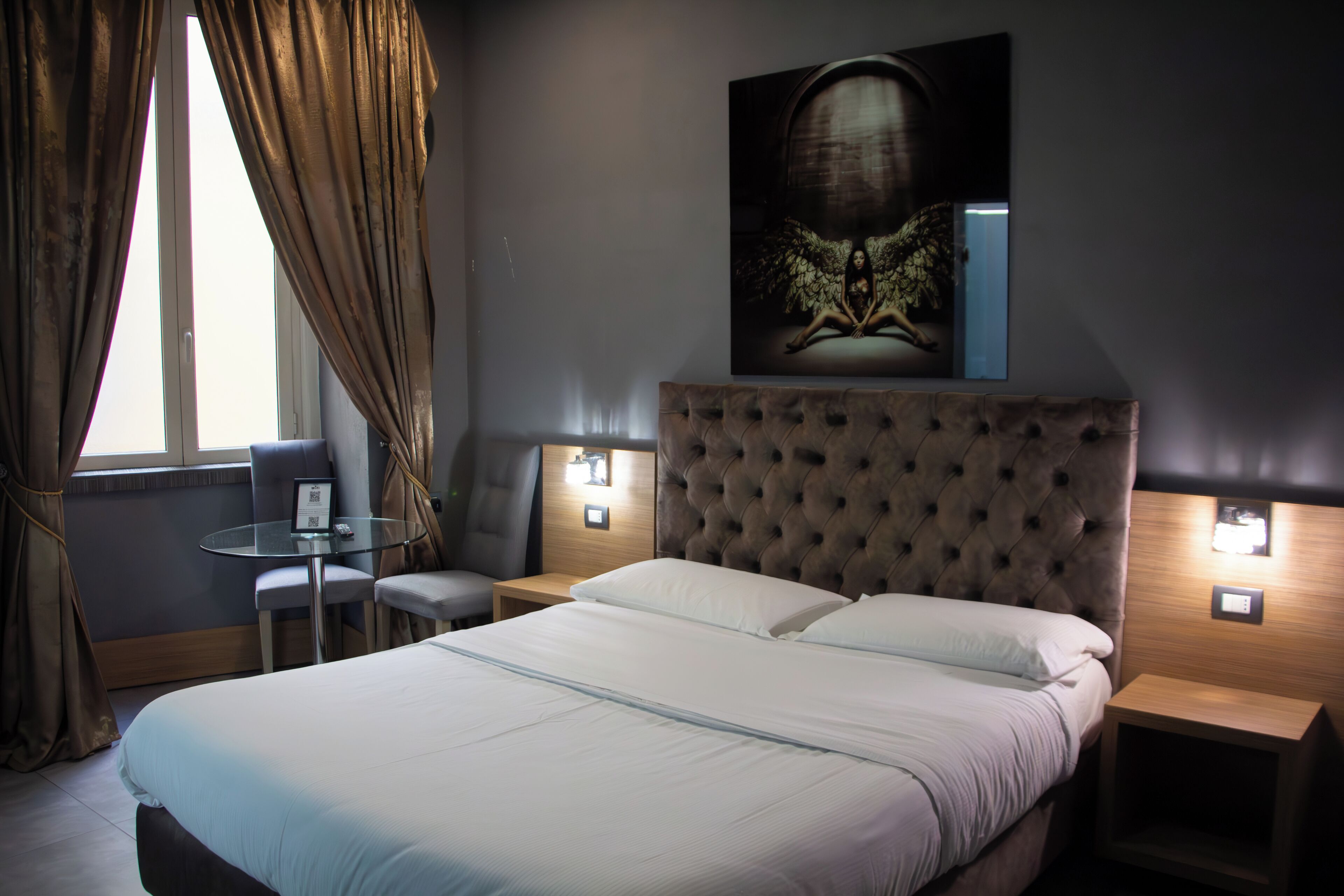 A hotel room with a large bed, two bedside tables, a small round table, and a chair. There is a large artwork on the wall above the bed.