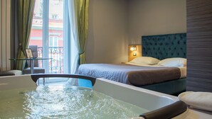 A hotel room with a bathtub, a bed with a blue headboard, and a view of a balcony with a railing. - Umberto Primo Suite e Spa (Naples)