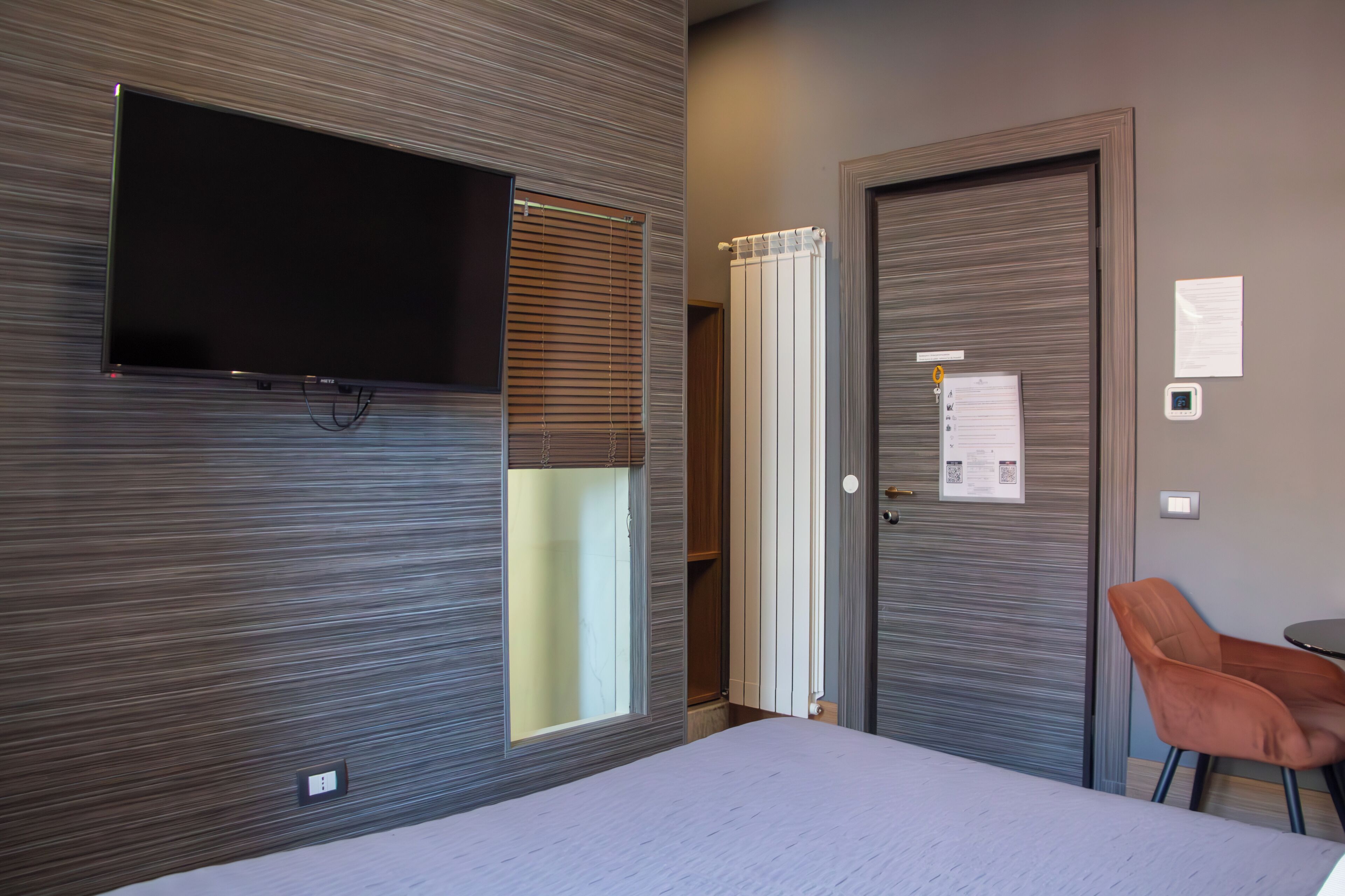Una habitación de hotel con una cama, un televisor montado en la pared, una puerta con un papel adherido y una silla.