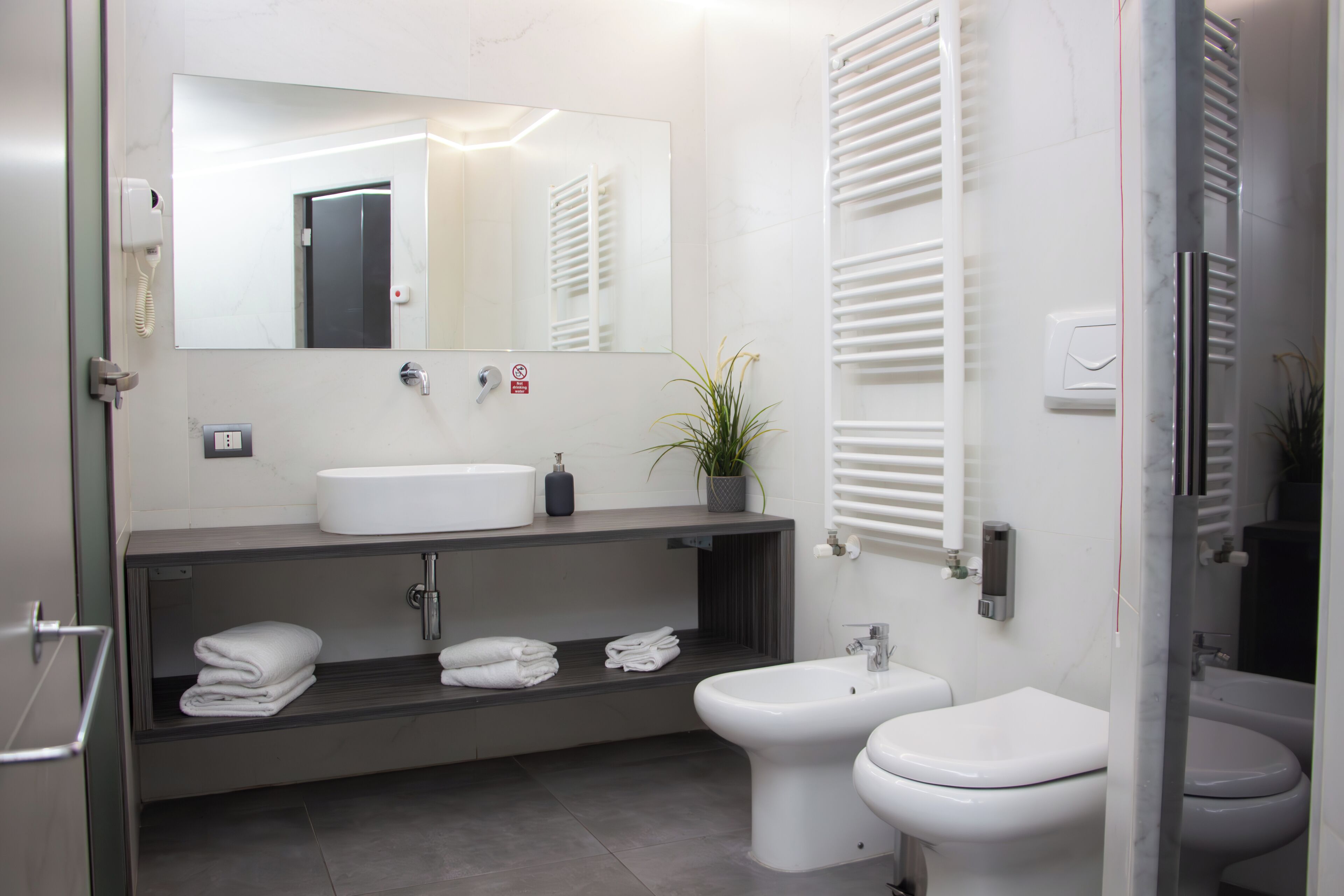 Une salle de bain moderne avec un lavabo blanc, un bidet et des toilettes. Un miroir est placé au-dessus du lavabo et un porte-serviettes en dessous.
