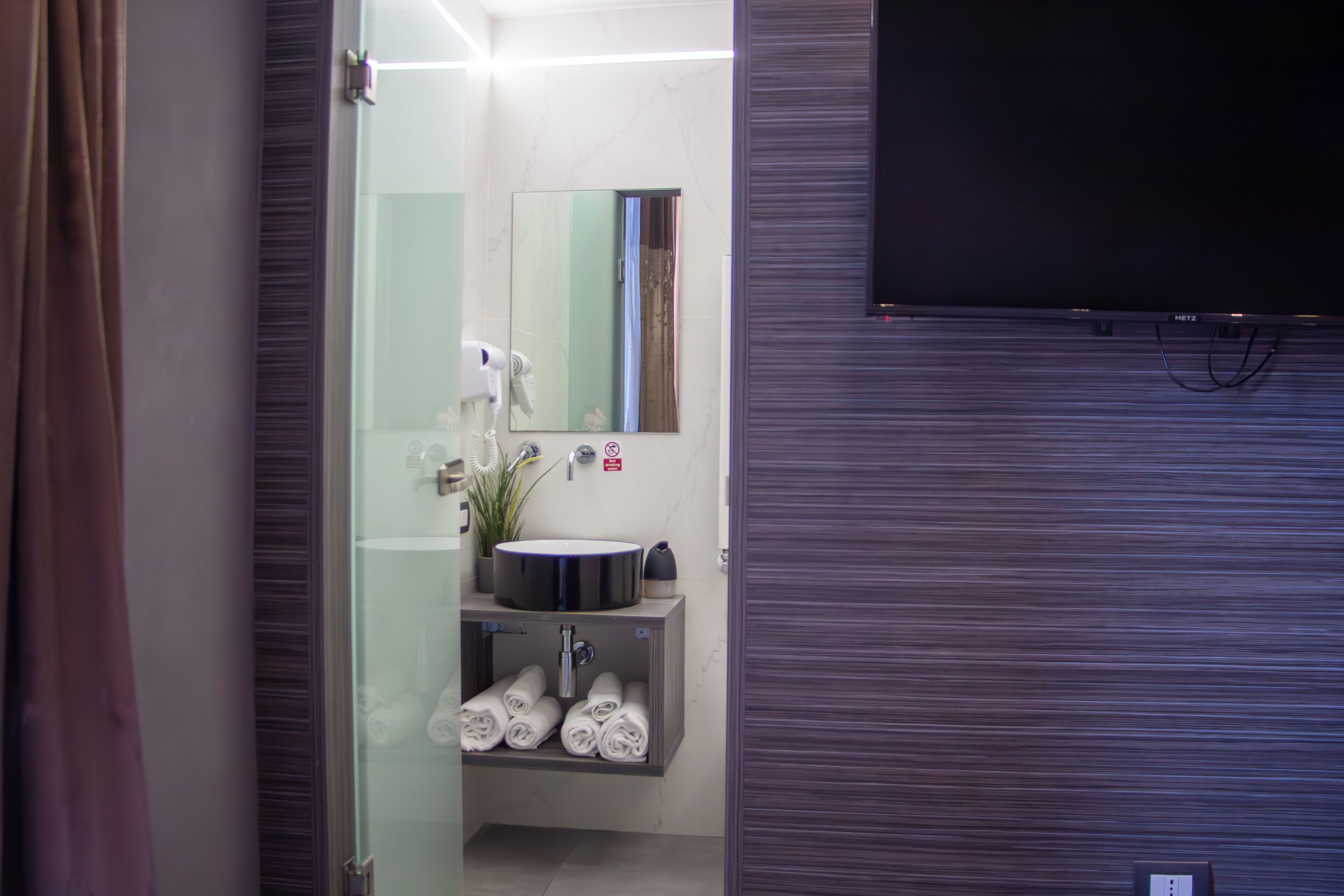 A modern bathroom with a glass shower enclosure, a sink with a potted plant, and neatly rolled towels.