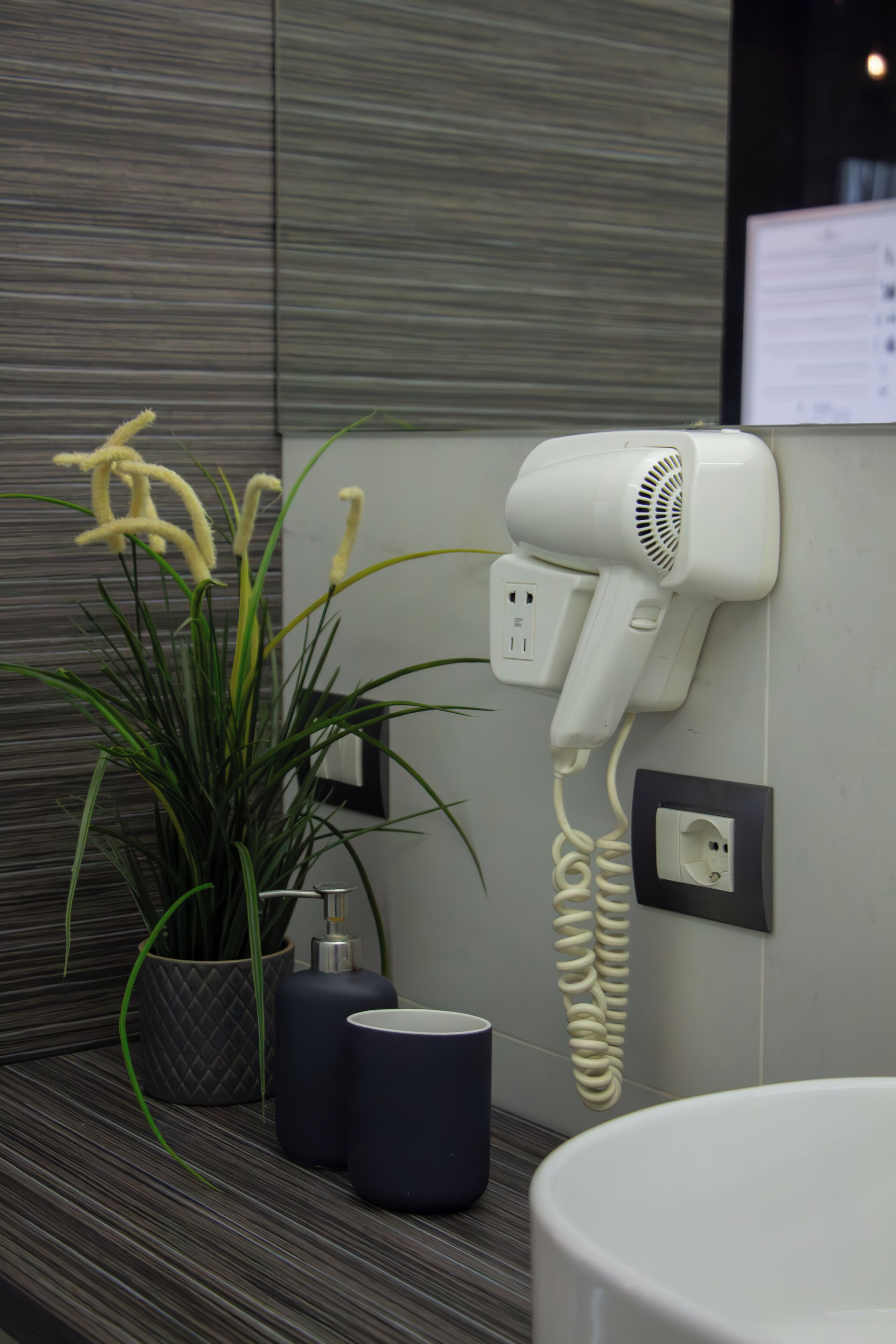 A hair dryer mounted on a wall, a potted plant, and two dark-colored cups on a striped countertop.