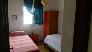 A bedroom with two beds, a window with curtains, a bedside table with a lamp, and a wardrobe.