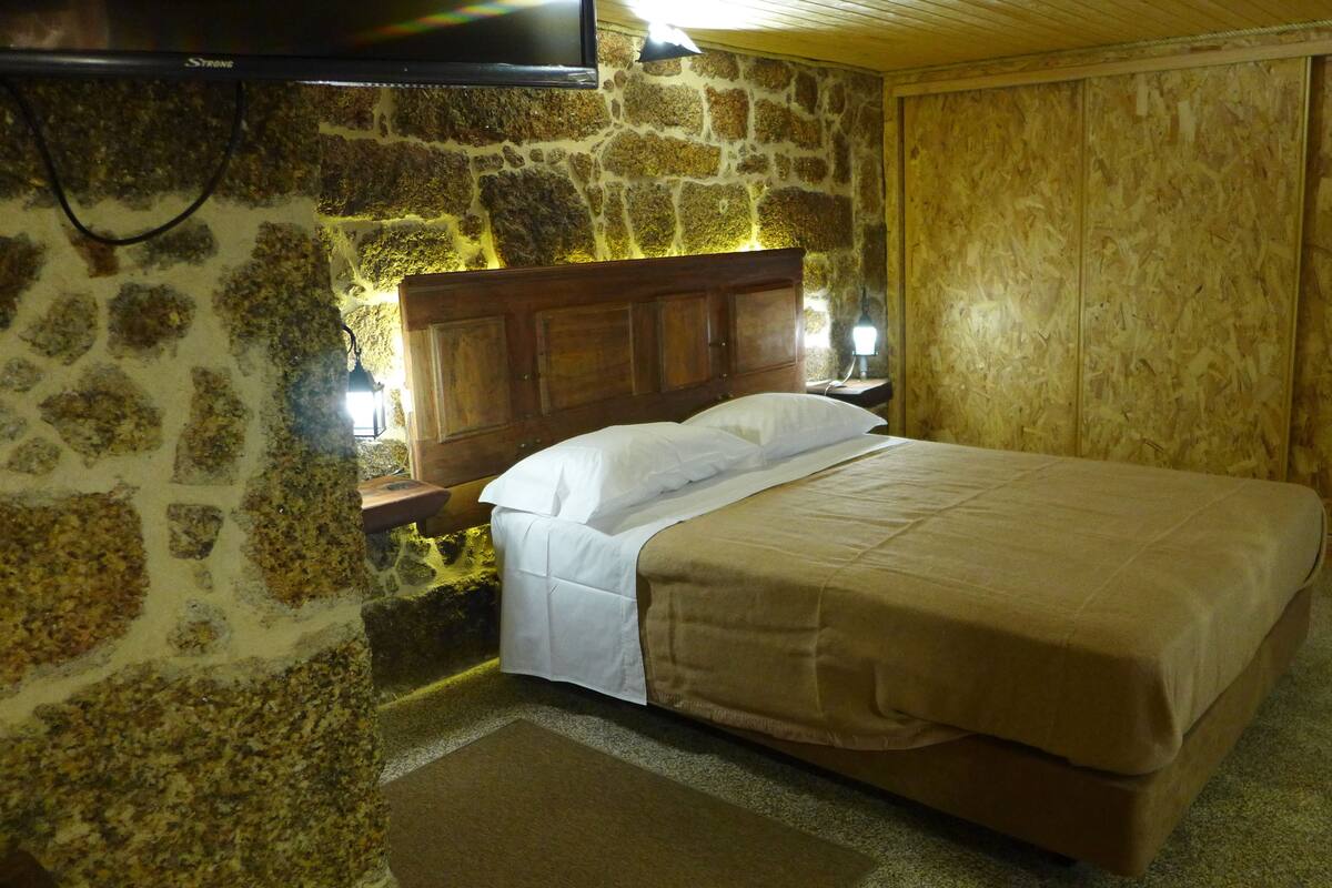 A bedroom with a stone wall, wooden headboard, a bed with white and brown bedding, and a TV mounted on the wall.