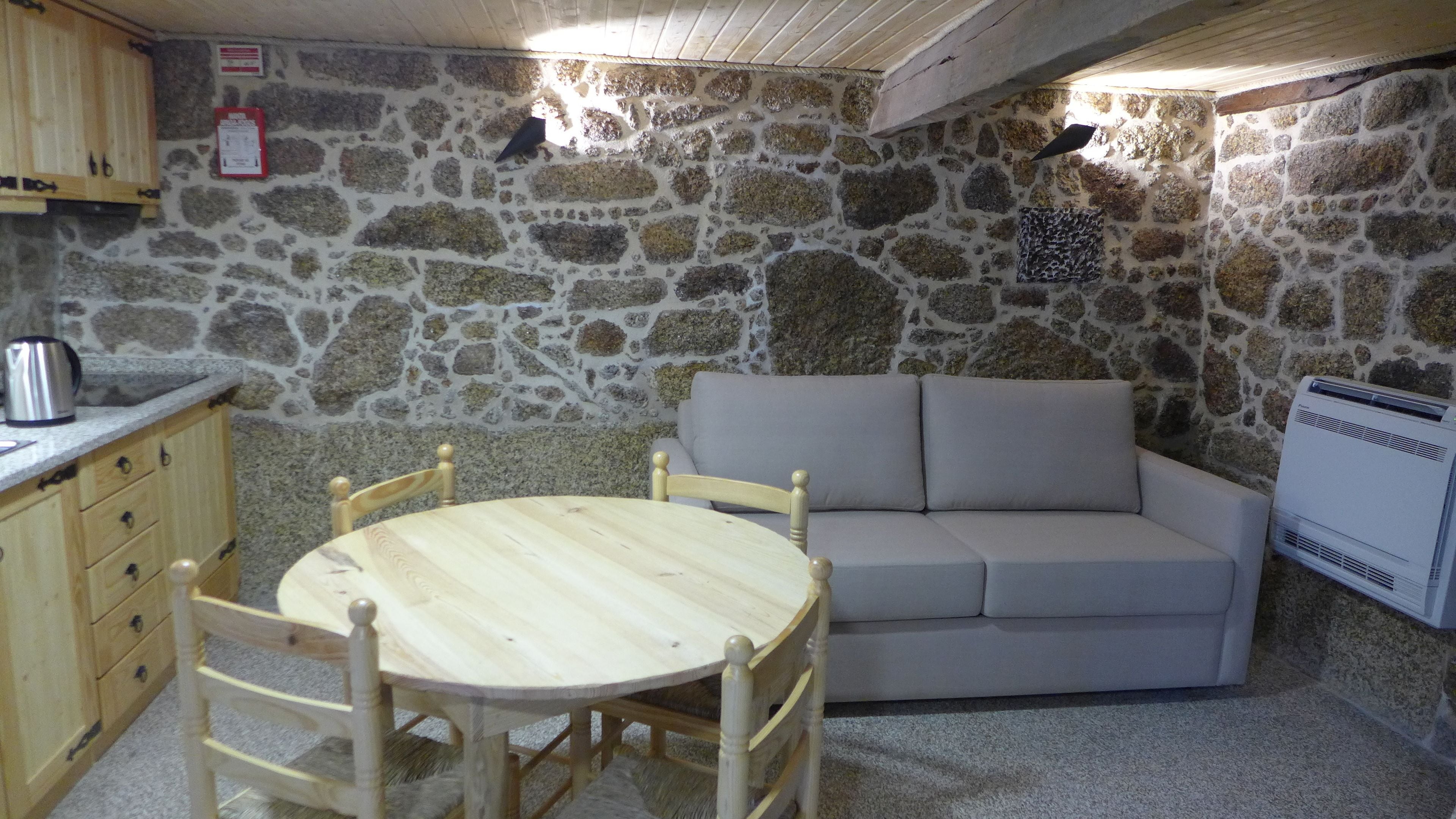 A stone-walled room with a round wooden table and chairs, a beige sofa, a kettle, and an air conditioner.