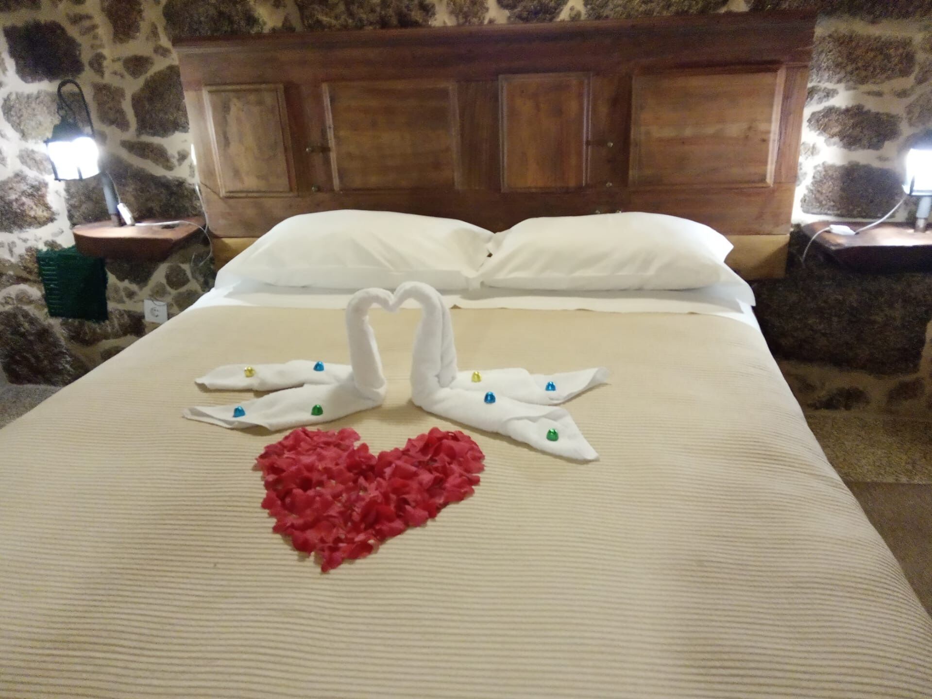 A bed with a swan and heart towel art, stone walls, wooden headboard, and two bedside lamps.
