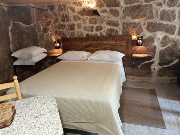A stone-walled bedroom with a wooden headboard, a bed with white linens, two bedside tables with lamps, and a small wooden chair.