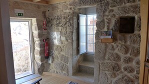 Una habitación con paredes de piedra, un extintor, un ventanal pequeño y una estantería de madera con libros.