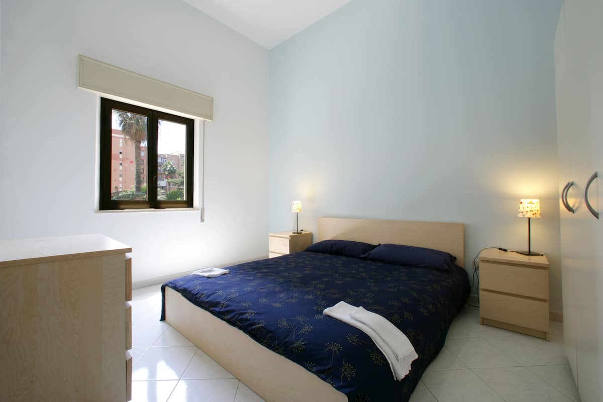 A bedroom with a bed, a dresser, two bedside tables with lamps, a window, and a view of buildings outside.