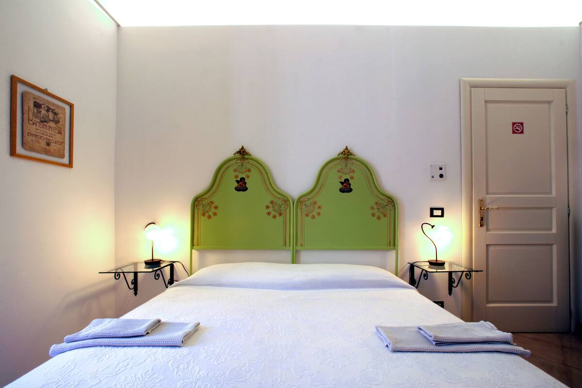 A hotel room with a double bed, two bedside tables with lamps, a door with a no-smoking sign, and a framed picture on the wall.