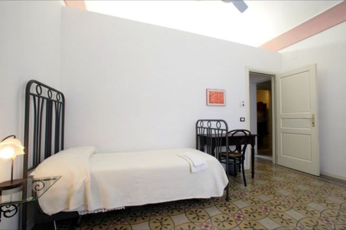 A small, well-lit bedroom with a single bed, a chair, a table, and a door leading to another room.