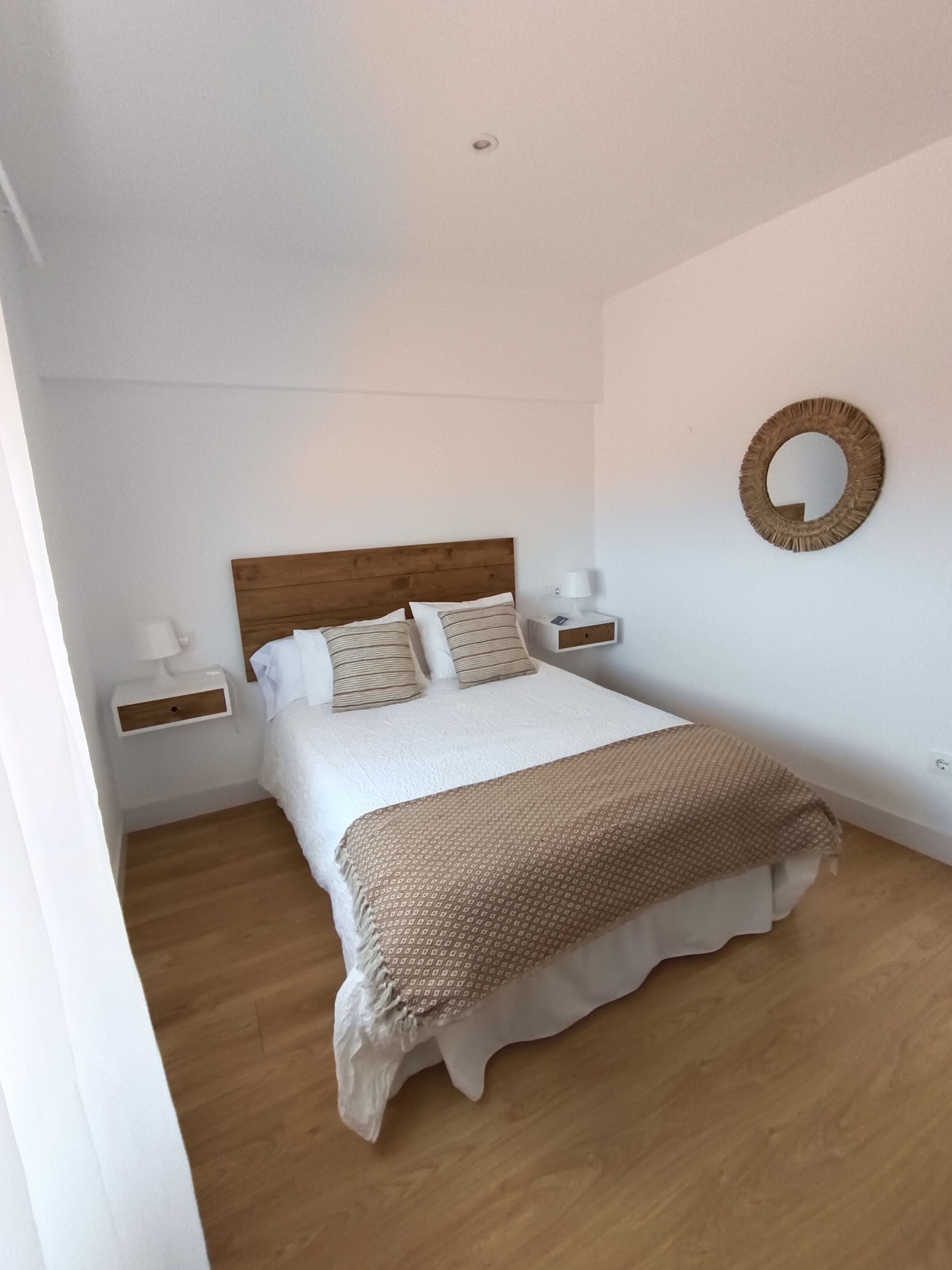A bedroom with a wooden headboard, a white bedspread, and a patterned throw blanket. There are two bedside tables with lamps and a wall-mounted mirror.