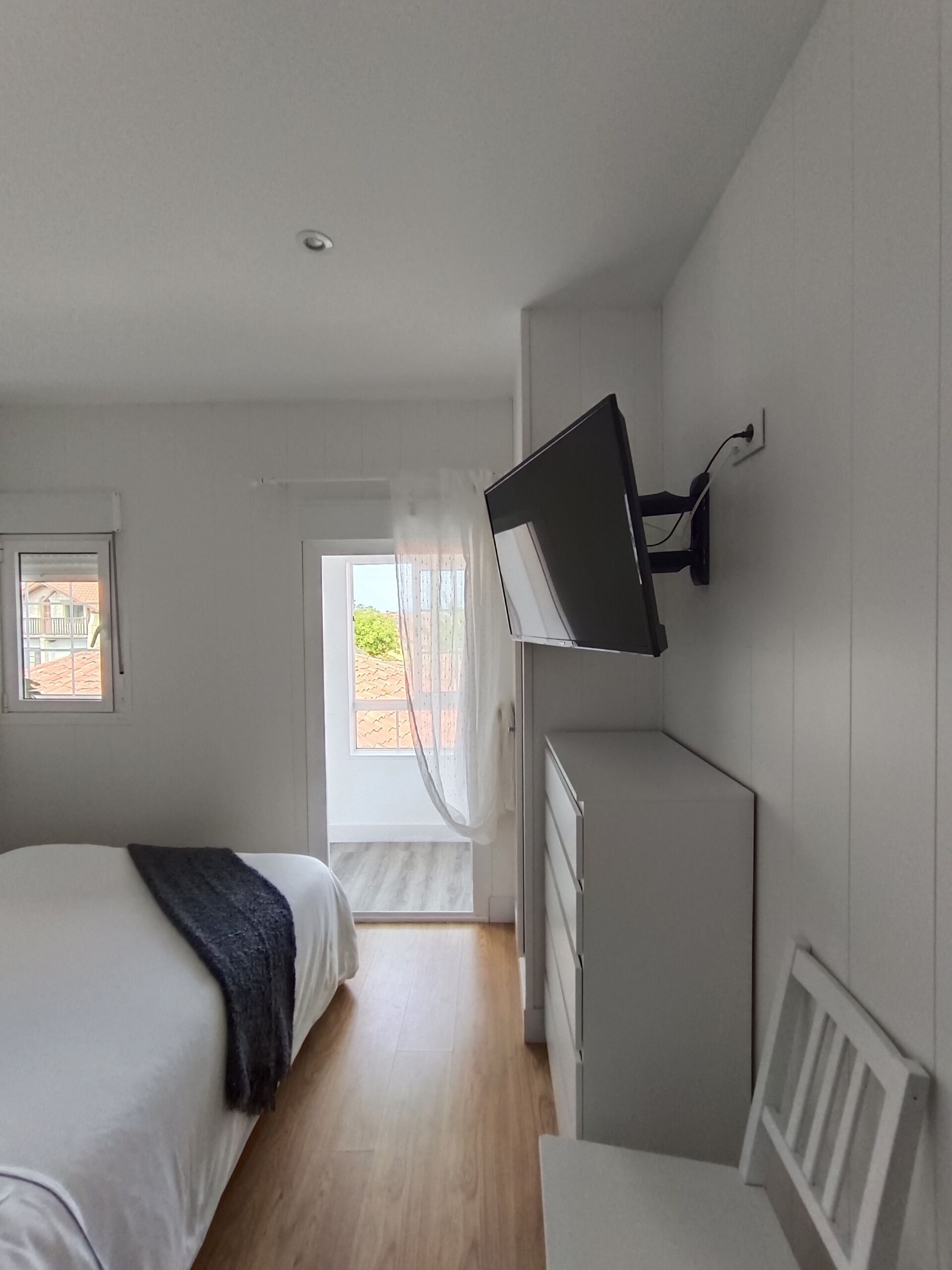A bedroom with a bed, a desk, a TV mounted on the wall, and a window with curtains.