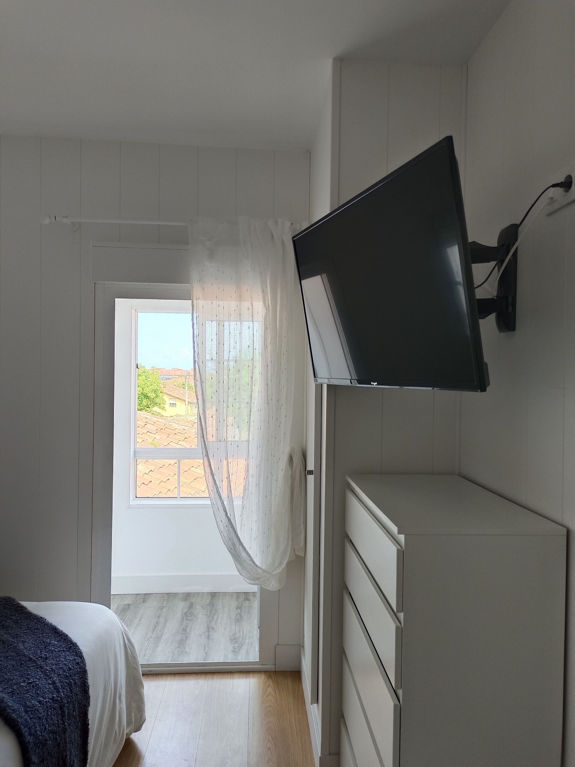 Un dormitorio con cama, televisor montado en la pared, ventana con cortina translúcida y piso de madera.