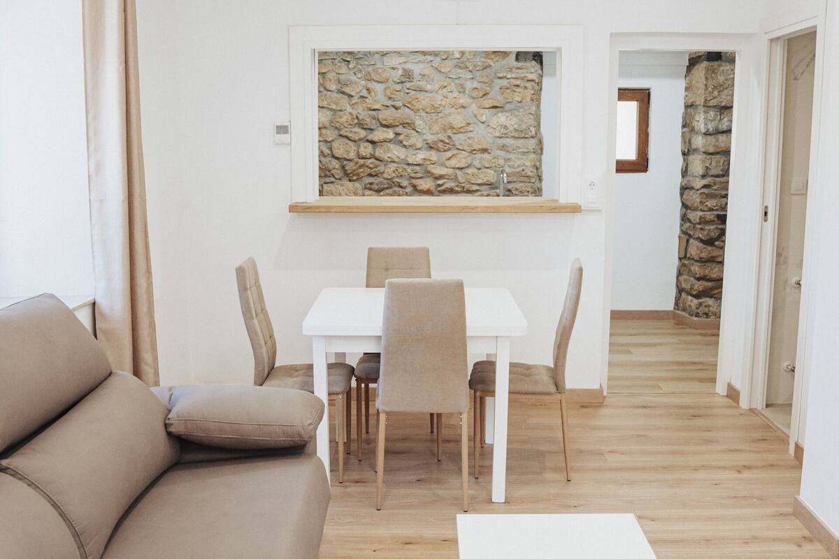 A modern living room with a beige sofa, a white coffee table, and a dining area with a white table and chairs. A stone wall and a wooden door are visible in the background.
