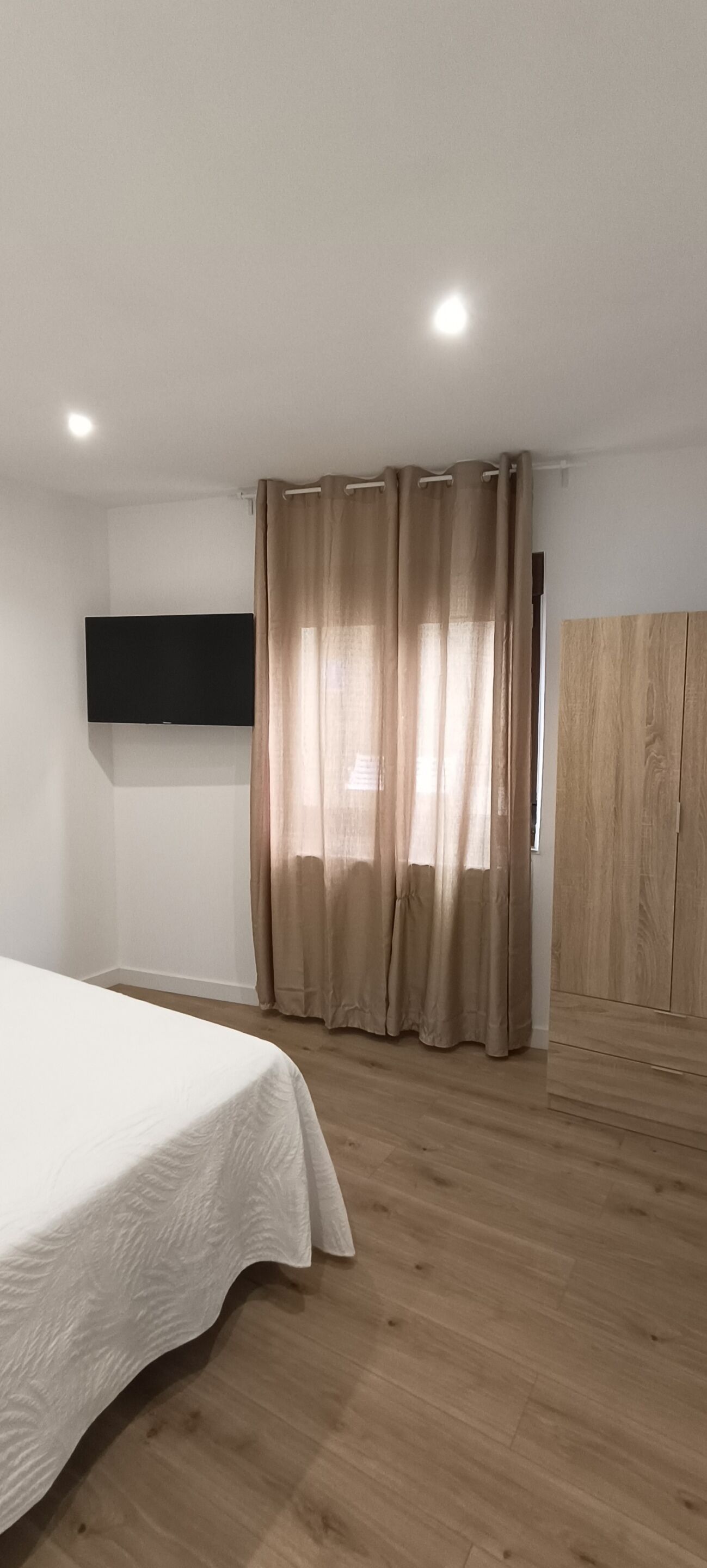 Un dormitorio con cama, piso de madera, televisor montado y ventana con cortinas.