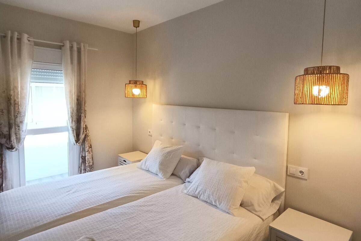A double bed with white linens and pillows, a bedside table, a nightstand, a window with curtains, and two wall-mounted lamps.