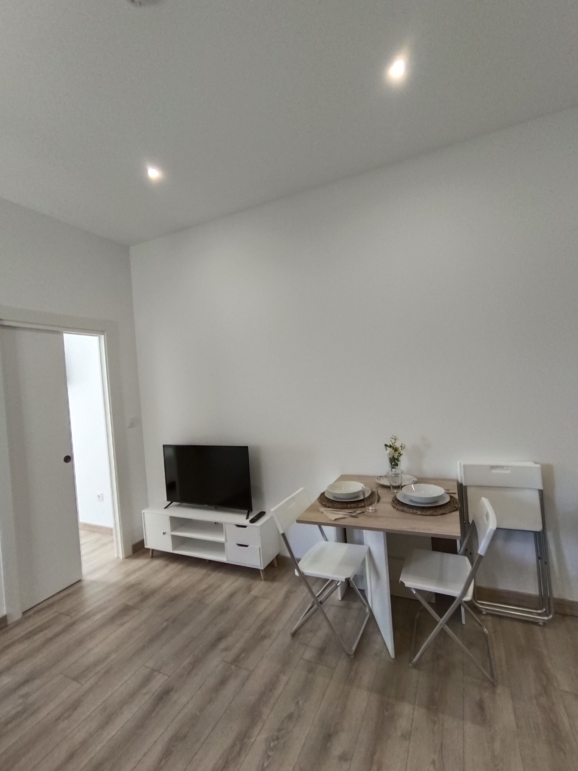 A modern, minimalist living room with a flat-screen TV on a white entertainment unit, a dining table with chairs, and a compact kitchenette.