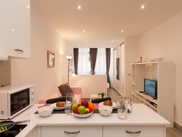 A modern kitchen with a microwave, a dining table set with fruits and a glass of water, a sofa, and a TV in the background.