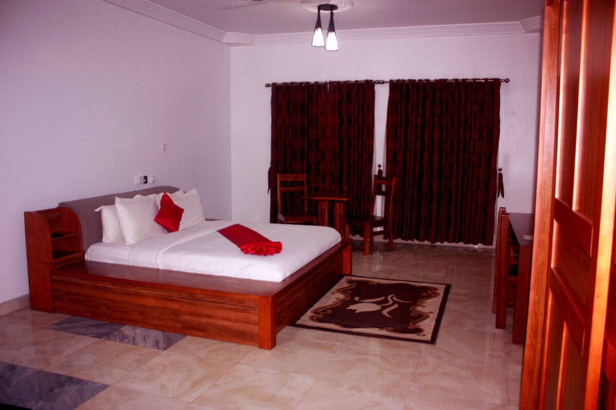 A bedroom with a wooden bed, white bedding, a red throw, a wooden nightstand, a chair, a desk, and a patterned rug.