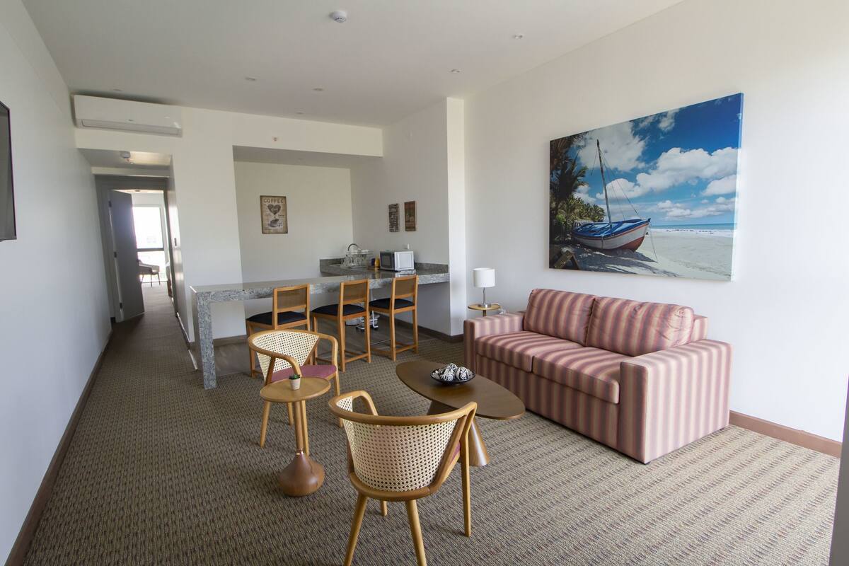 A hotel room with a living area, a dining area, and a kitchen. There is a painting of a boat on the wall.