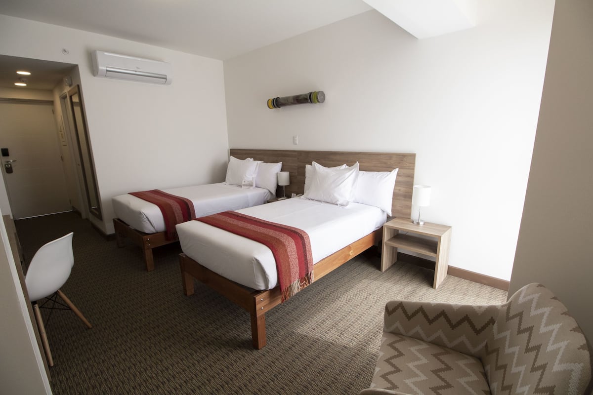 A hotel room with two single beds, a small desk, a chair, and a large window.