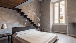 A modern bedroom with a large bed, a wooden ceiling, a staircase, and a window with a view of buildings. - RSH Ara Pacis Enchanting Apartment (Rome)