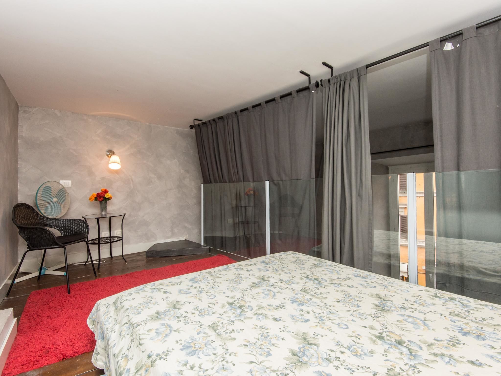 A bedroom with a bed, a chair, a small table with flowers, a wall-mounted light, and a sliding glass door.