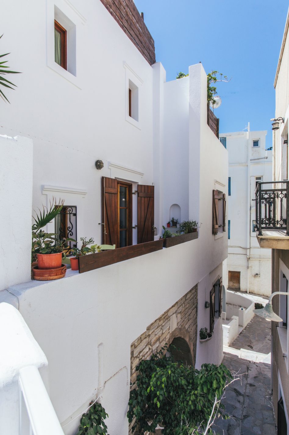 A narrow, white-walled building with a stone base, wooden shutters, and potted plants on the balcony.