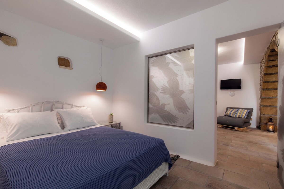 Un dormitorio con una cama, un televisor montado en la pared y un ventanal con un cuadro de un pájaro.
