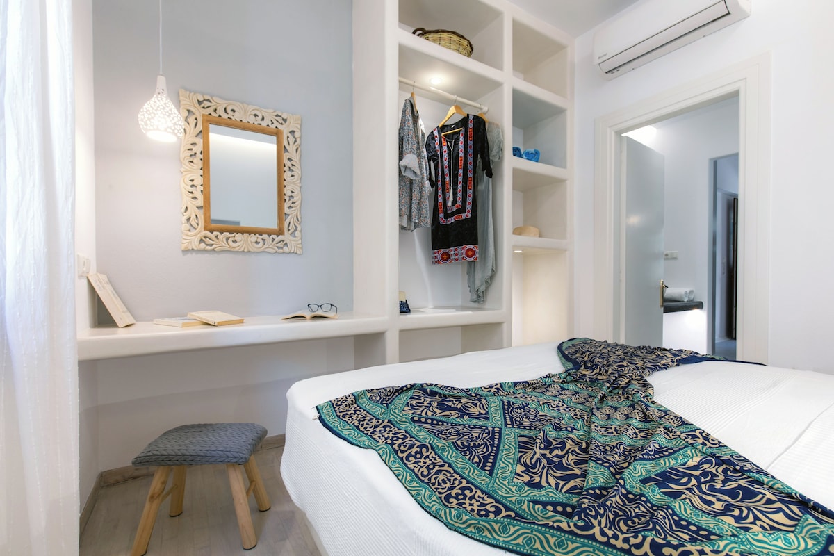 Un dormitorio con cama, un taburete pequeño, un armario, un espejo y una lámpara colgante.