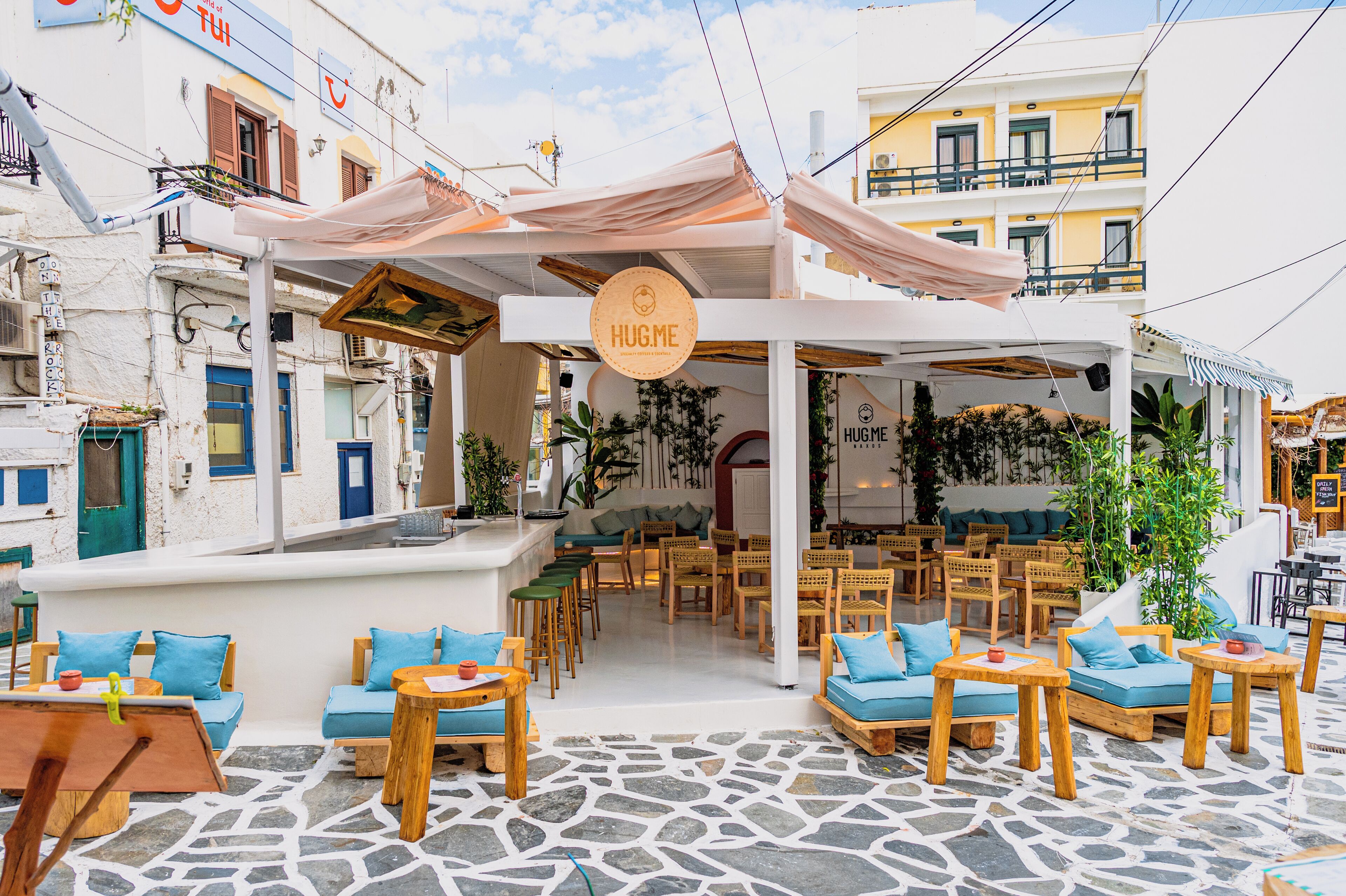 A cozy outdoor seating area with wooden furniture and a stone floor, surrounded by a white building with balconies and a 'HUG.ME' sign.