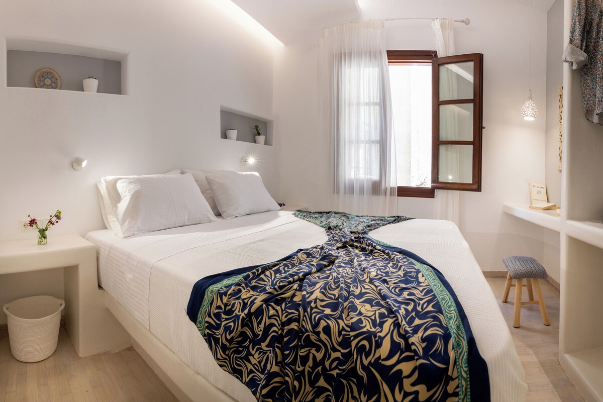 Un dormitorio con cama, mesitas de noche, una pequeña taburete, una ventana con cortinas y una estantería con objetos decorativos.