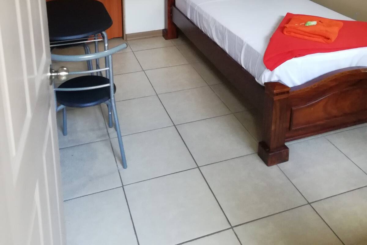 Ein kleiner, ordentlicher Raum mit einem hölzernen Bett, einem Sessel und einem kleinen Tisch.