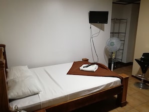 A single bed with a white mattress and brown headboard, a remote control on the bed, a wall-mounted TV, and a standing fan.
