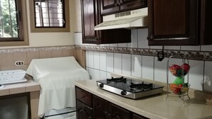 A kitchen with dark wooden cabinets, a gas stove, a white countertop, and a window with metal bars. - Hospedaje Yaldai (Turrialba)