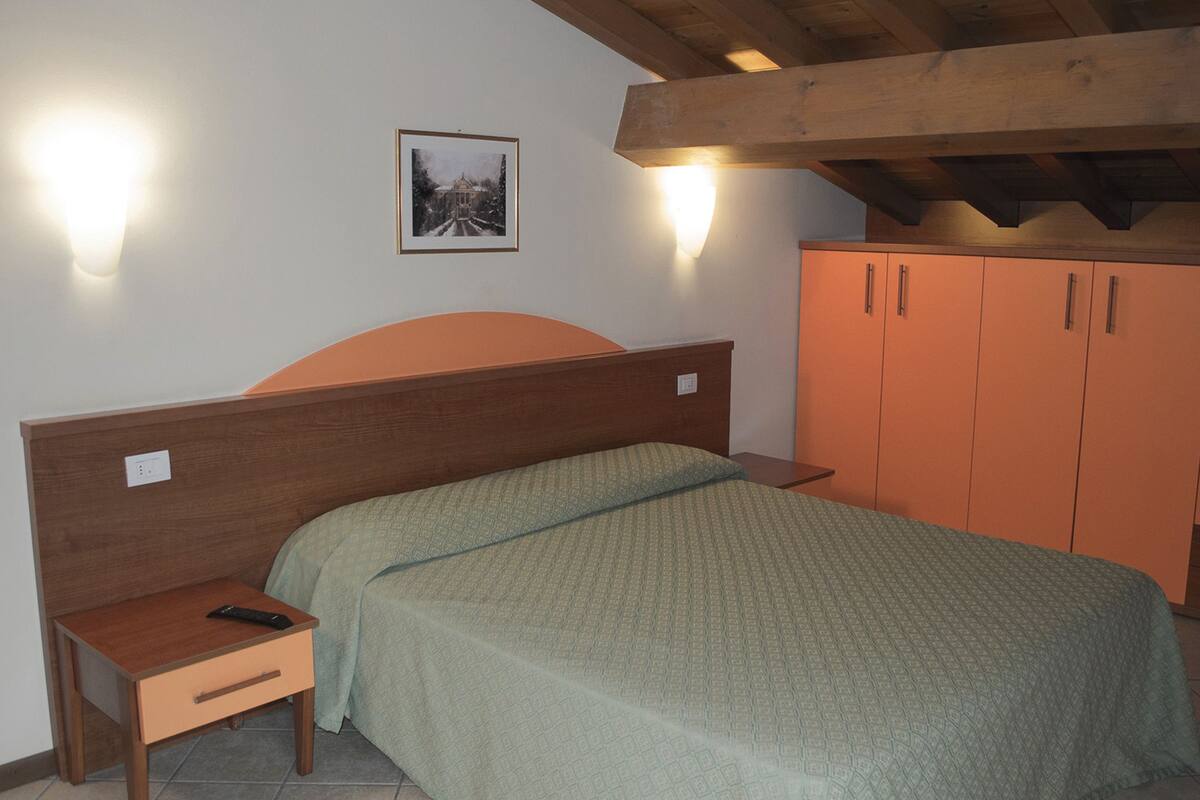 Une chambre à coucher avec une tête de lit en bois, un lit simple recouvert d’une couverture à motifs verts, une table de chevet avec une télécommande, une armoire orange et un tableau encadré accroché au mur.