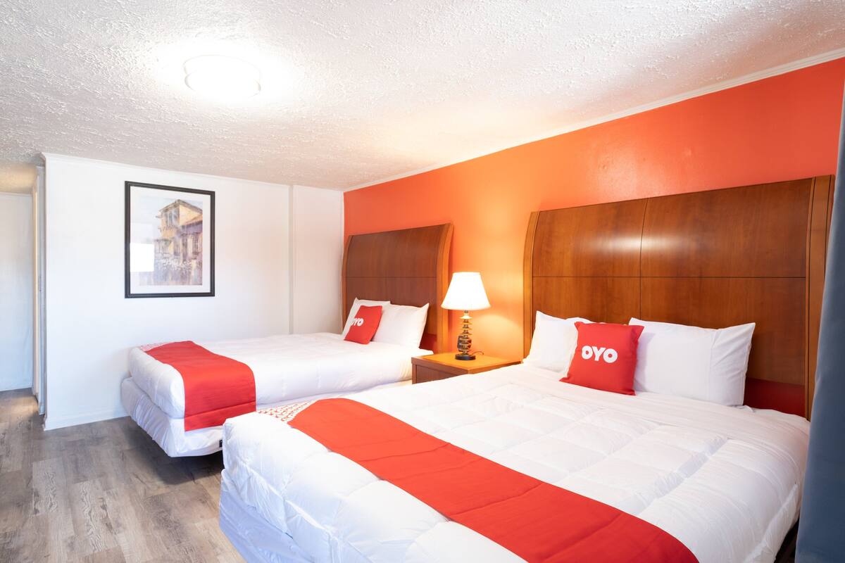 Une chambre d’hôtel avec deux lits, chacun agrémenté de draps blancs et de touches de couleur rouge, une tête de lit en bois, une table de chevet avec une lampe et un tableau encadré au mur.